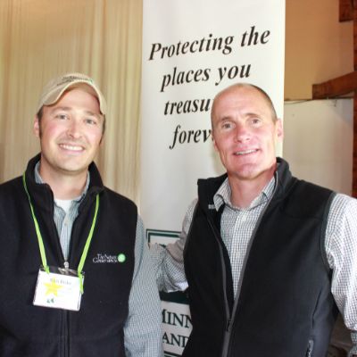 Two men smile into the camera posing for a photo, standing in front of a sign reading "Protecting the places you treasure...forever."