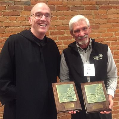 Two men standing in front of a brick wall smiling, holding awards plaques
