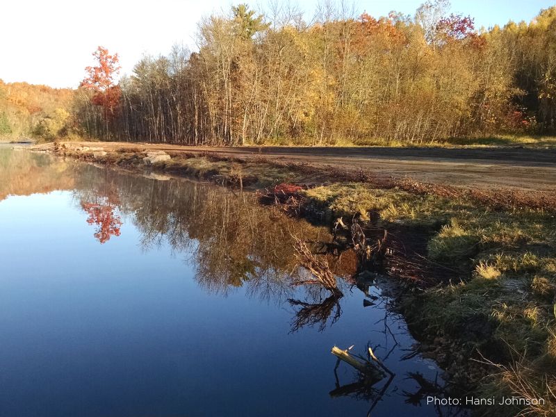 A photo of a natural river shoreline with calm, glassy water and fall foliage trees in the background.