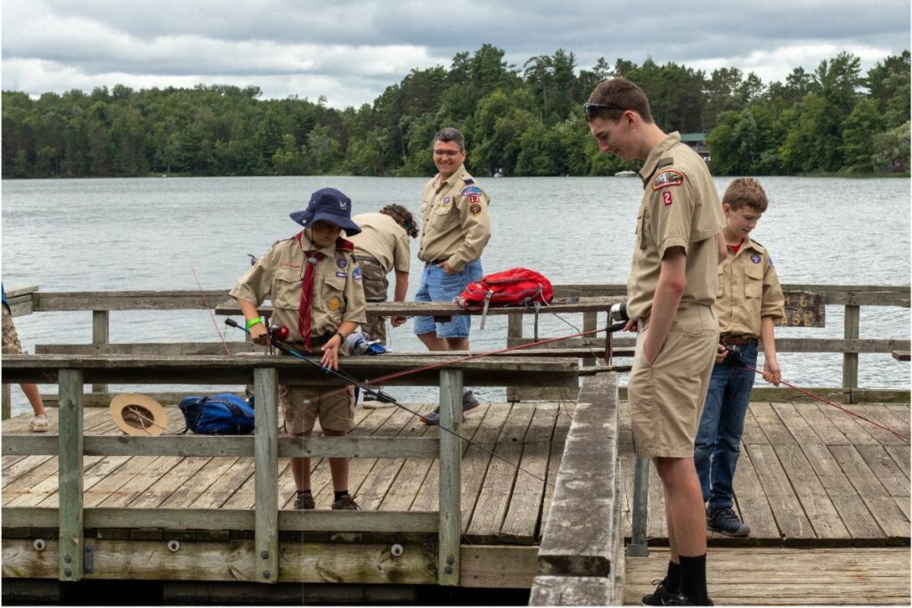 Several young men wearing Boy Scout uniforms stand on a wood dock with fishing poles