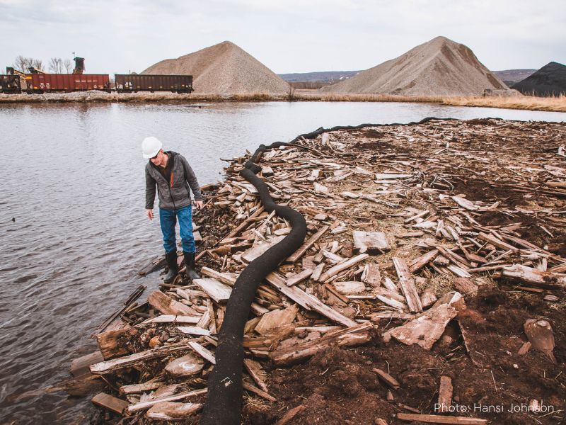 A photo of a man in a hard hat and jeans walking along a shore that is layered in drying sawmill debris wood, with an industrial site and train cars in the background.
