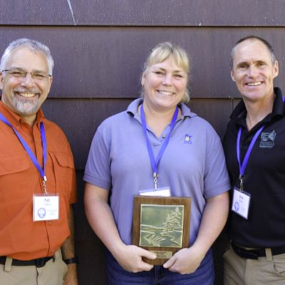 Three people pose for a photo, smiling, the middle person holds an award plaque