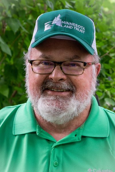 A man with a bear, glasses, a green shirt and cap smiles into the camera