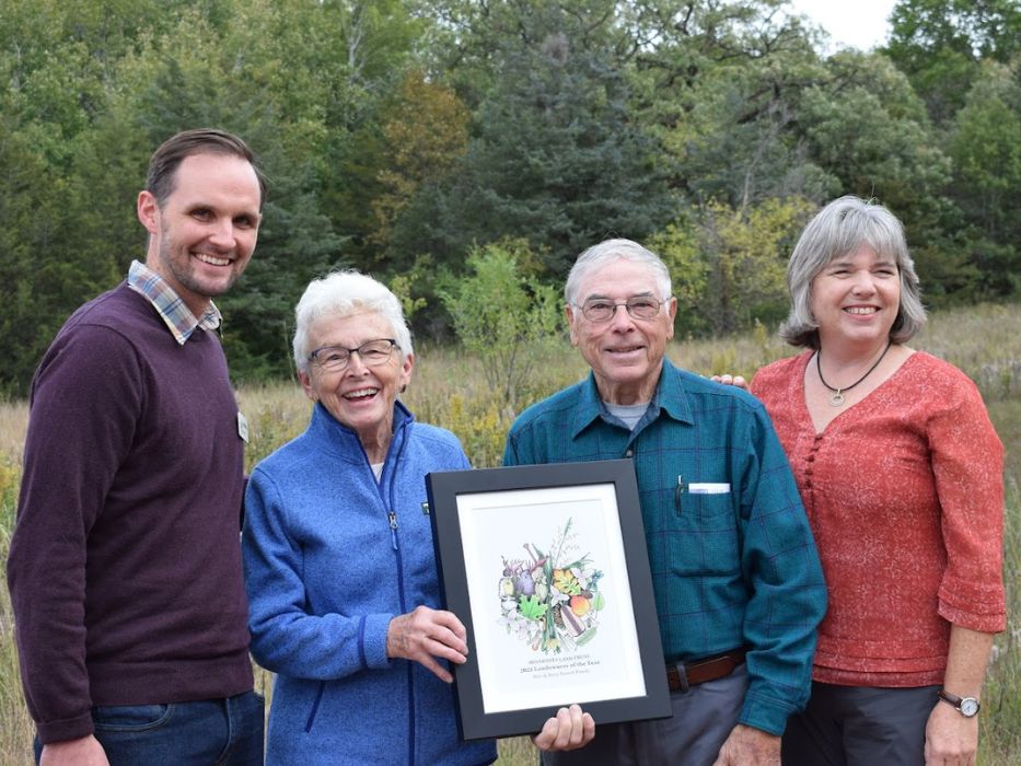 Minnesota Land Trust's Landowner of the Year 2024 award ceremony. Four individuals, including the recipients, are smiling and holding a framed award. They stand outdoors in front of a scenic natural backdrop.