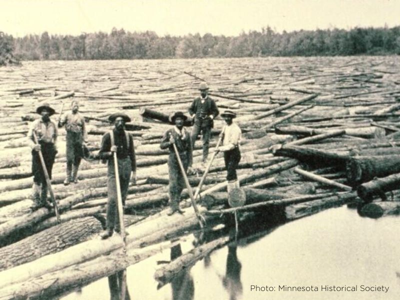A historic black and white photo of loggers (men) wearing hats and holding poles standing on tree trunks in the water during a "log roll".