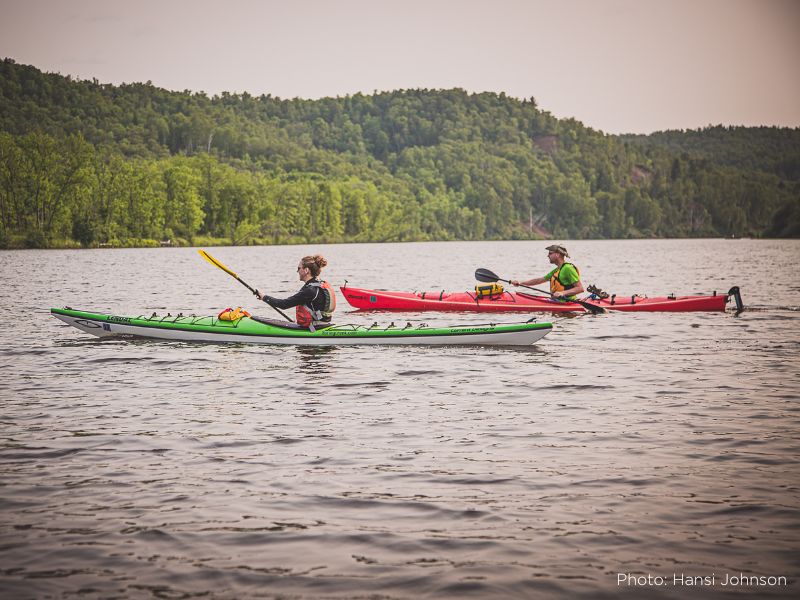 A photo of two people paddling kayaks in a river with a lush, green background of evergreen trees.