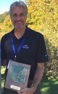 A man standing in front of a beautiful fall landscape poses for a photo, smiling, holding an award plaque