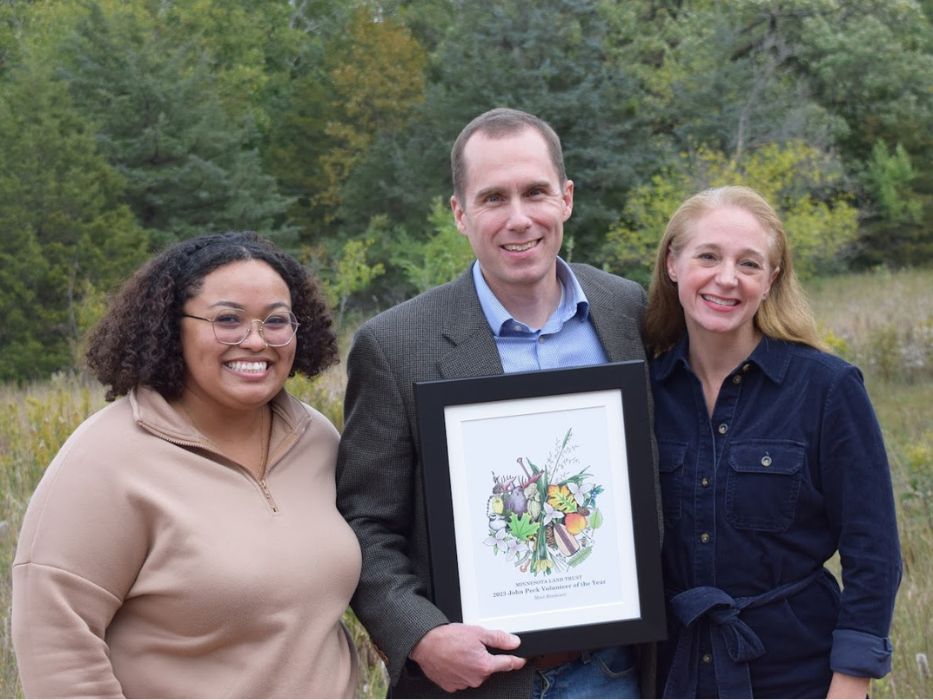Minnesota Land Trust's Volunteer of the Year 2024 award ceremony. Three people, including the award recipient Matt Bredesen, are standing outdoors with trees in the background, smiling and holding a framed award.